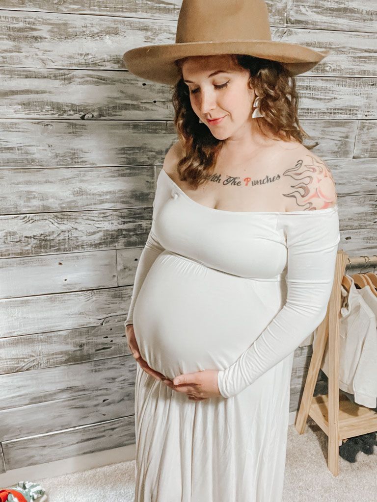 pregnant woman standing in front of a white washed wood wall wearing a white dress and brown hat looking down at her baby for about me blog page